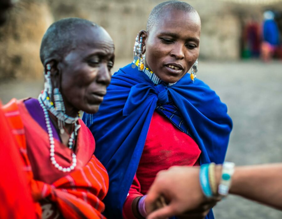 Two Maasai women adorned in vibrant traditional attire and beaded jewelry, engaged in conversation, with a blurred background of their community.